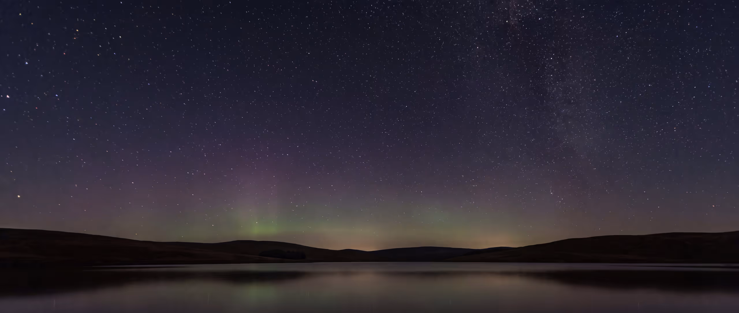A wide imagined night panorama with faint green and violet light over dark hills and still water beneath a dense field of stars.