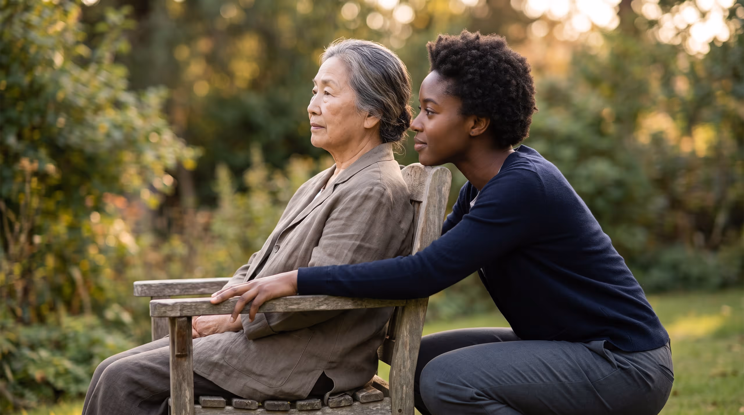 An elderly woman seated on a garden bench in late afternoon light, and a younger woman crouched beside her, listening.