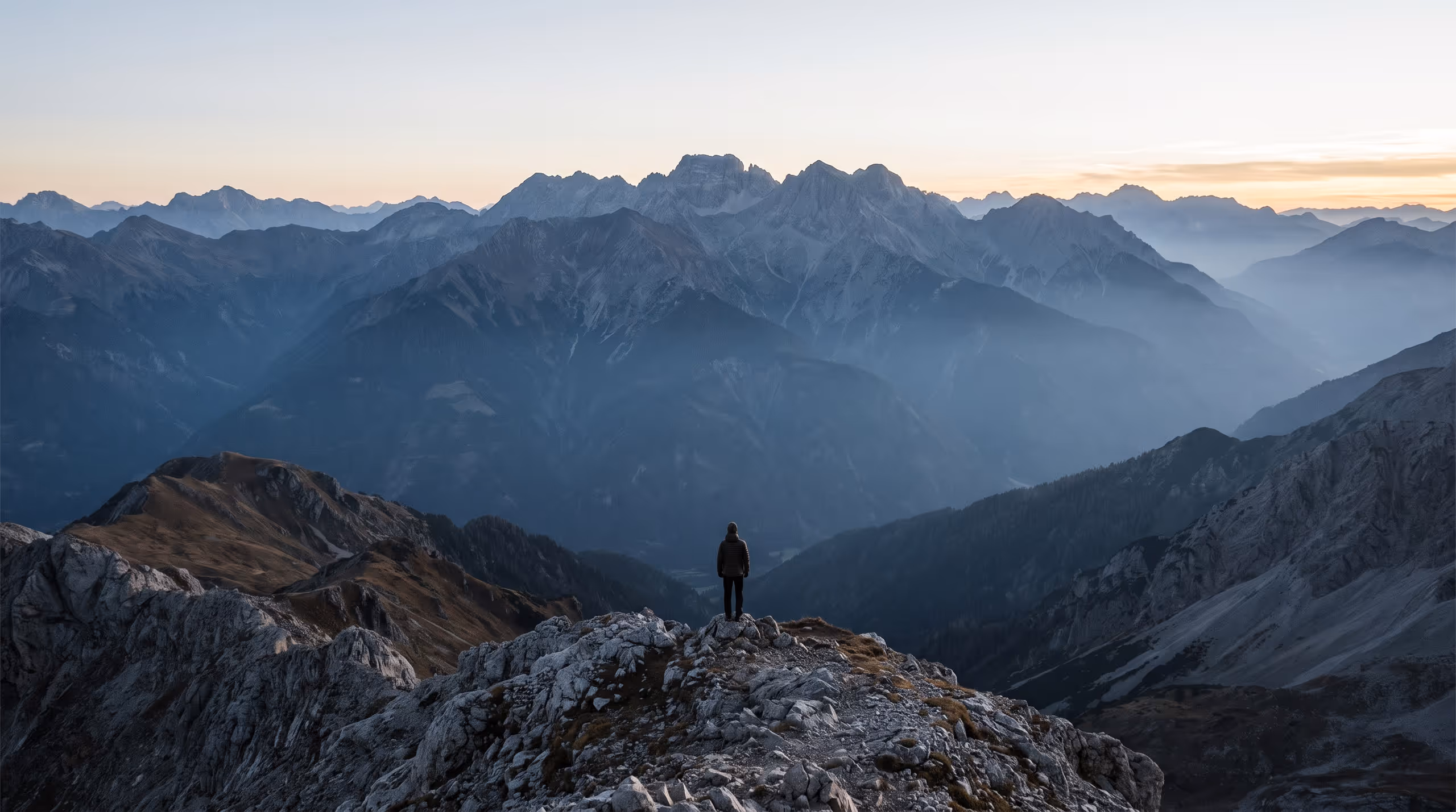 A vast mountain range at dawn in receding blue planes, with a single very small figure standing on a near ridge.