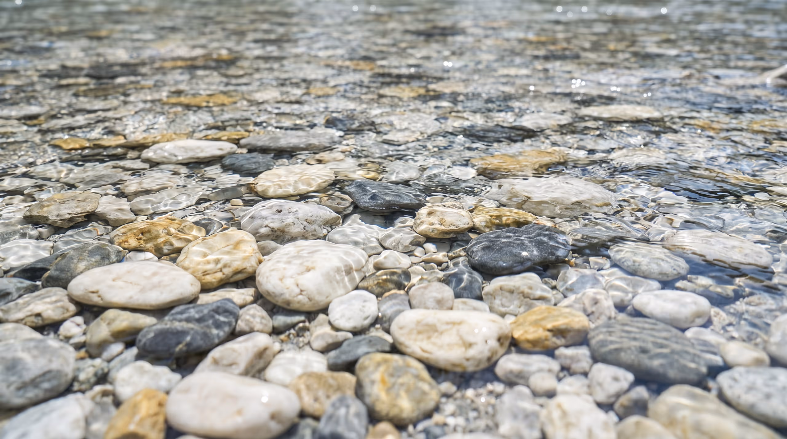 Clear shallow water running over pale rounded pebbles in bright daylight, every stone sharply visible through the surface.
