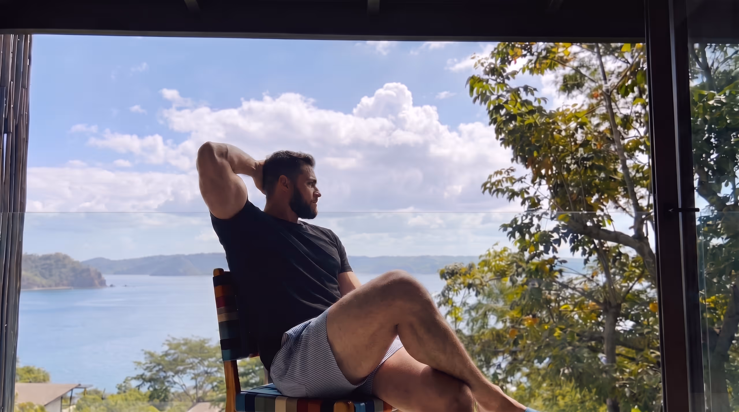 A person reclining in a low chair on a shaded terrace, one arm behind their head, looking out over a calm bay to distant blue headlands.