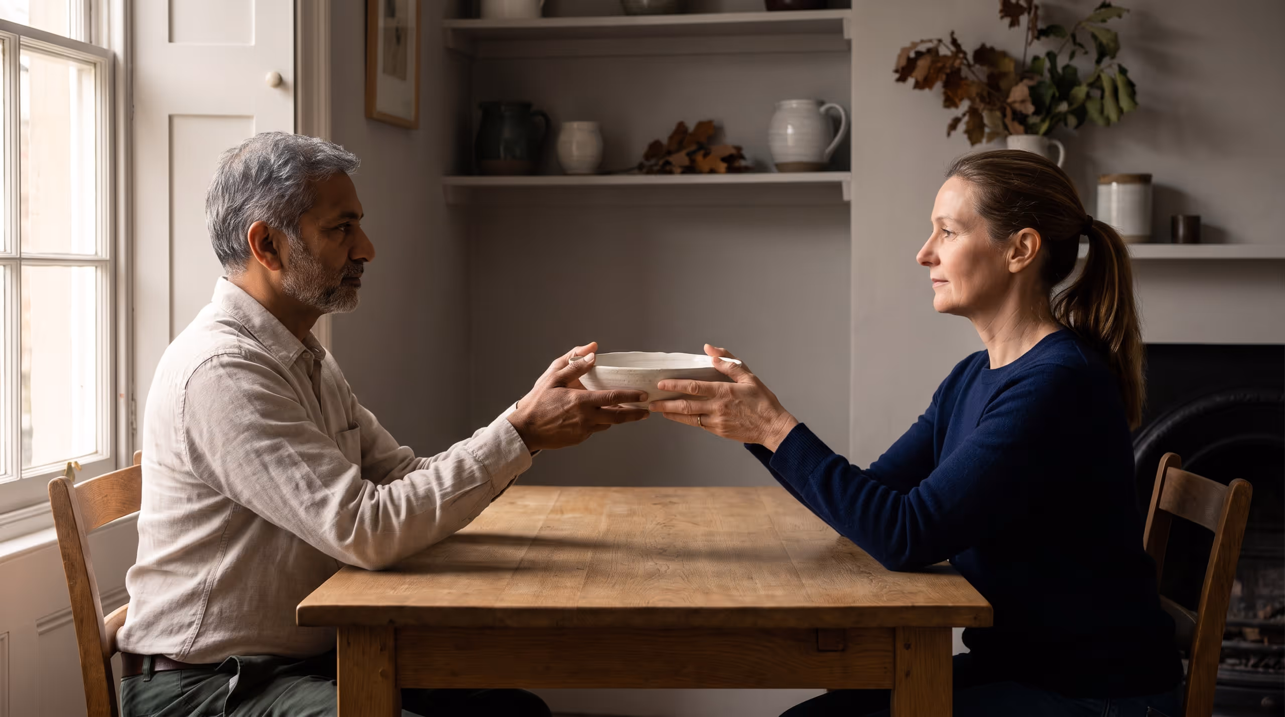 Two people seated facing each other at a plain table, both pairs of hands together on a shallow bowl held between them, mid-exchange.