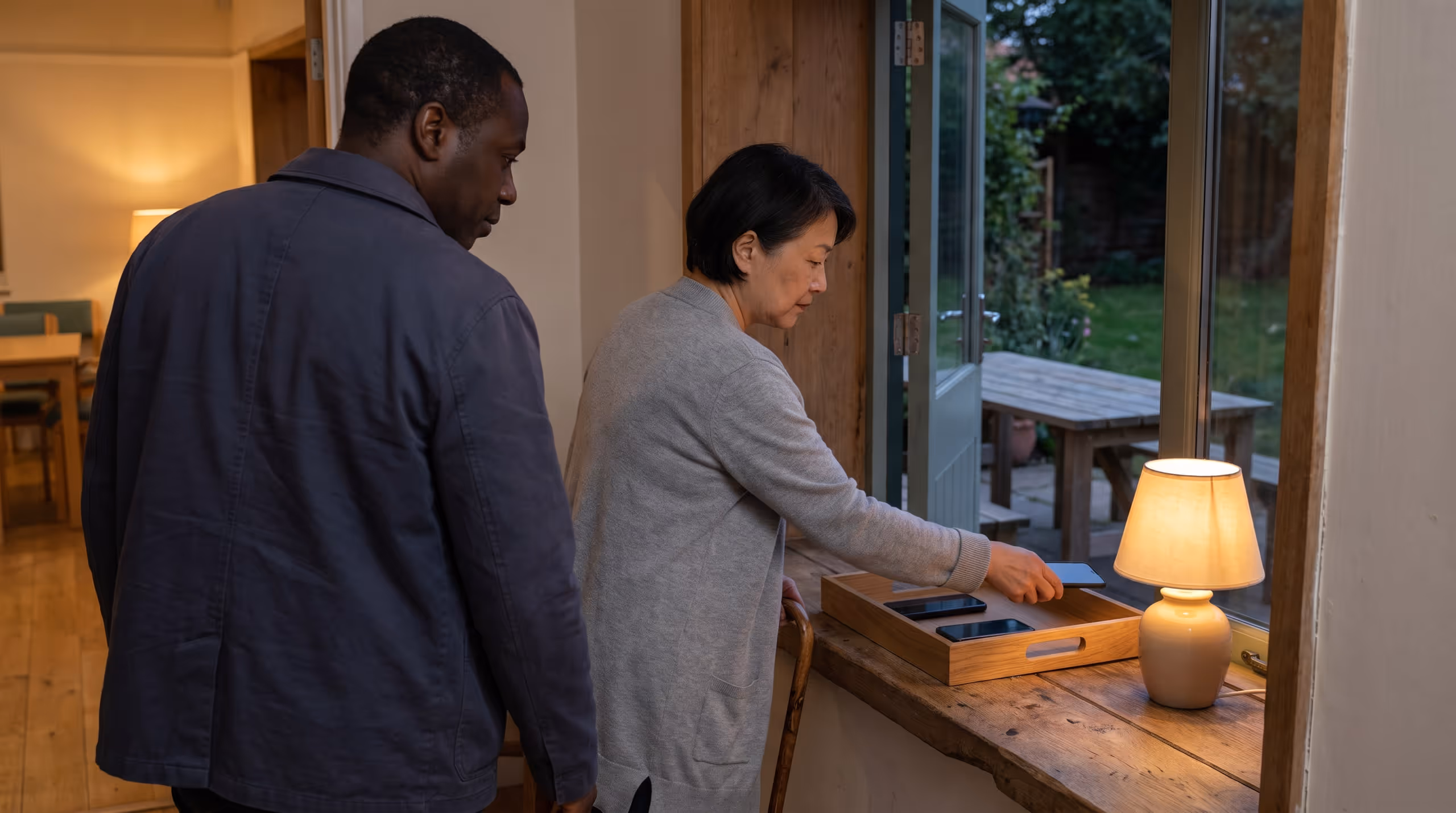 Two adults at a timber threshold placing their phones face down in a shallow tray before stepping into a garden at dusk.
